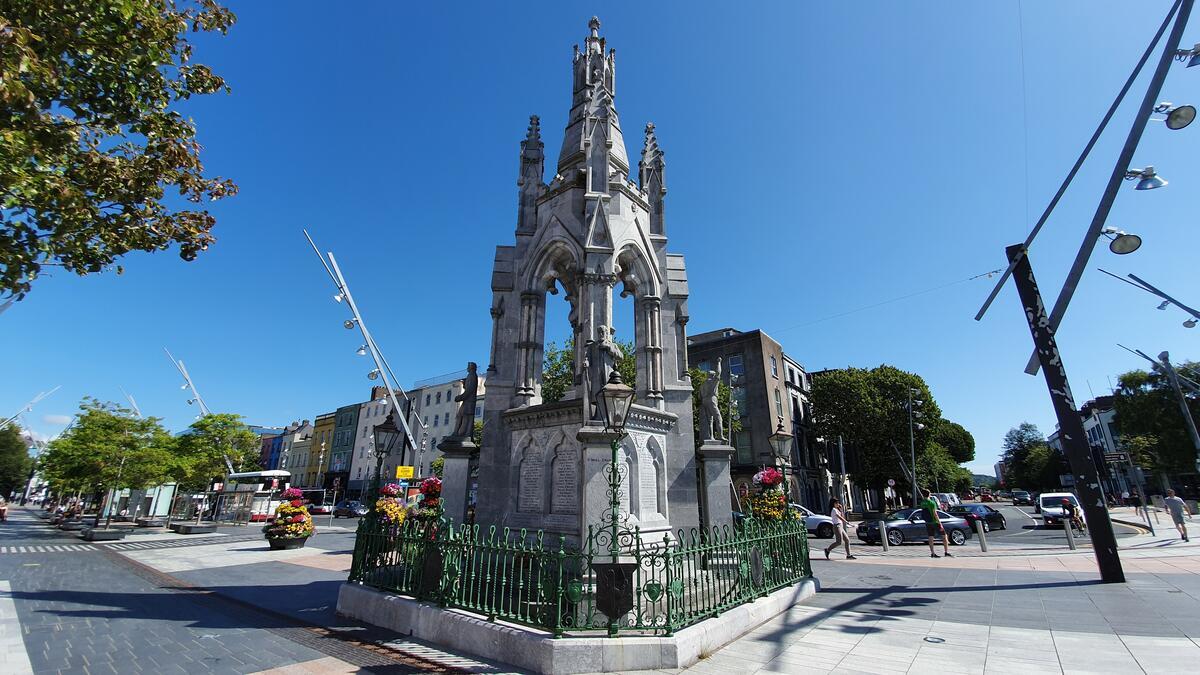 The National Monument on Grand Parade in Cork city, with flower baskets, passing traffic and a clear blue sky above the city centre.